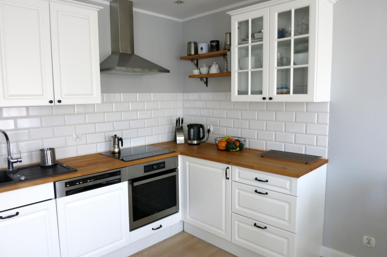 Jasna kuchnia z białymi szafkami, drewnianym blatem, płytkami w stylu metro i srebrnym okapem; na blacie czajnik, owoce i kawiarka.