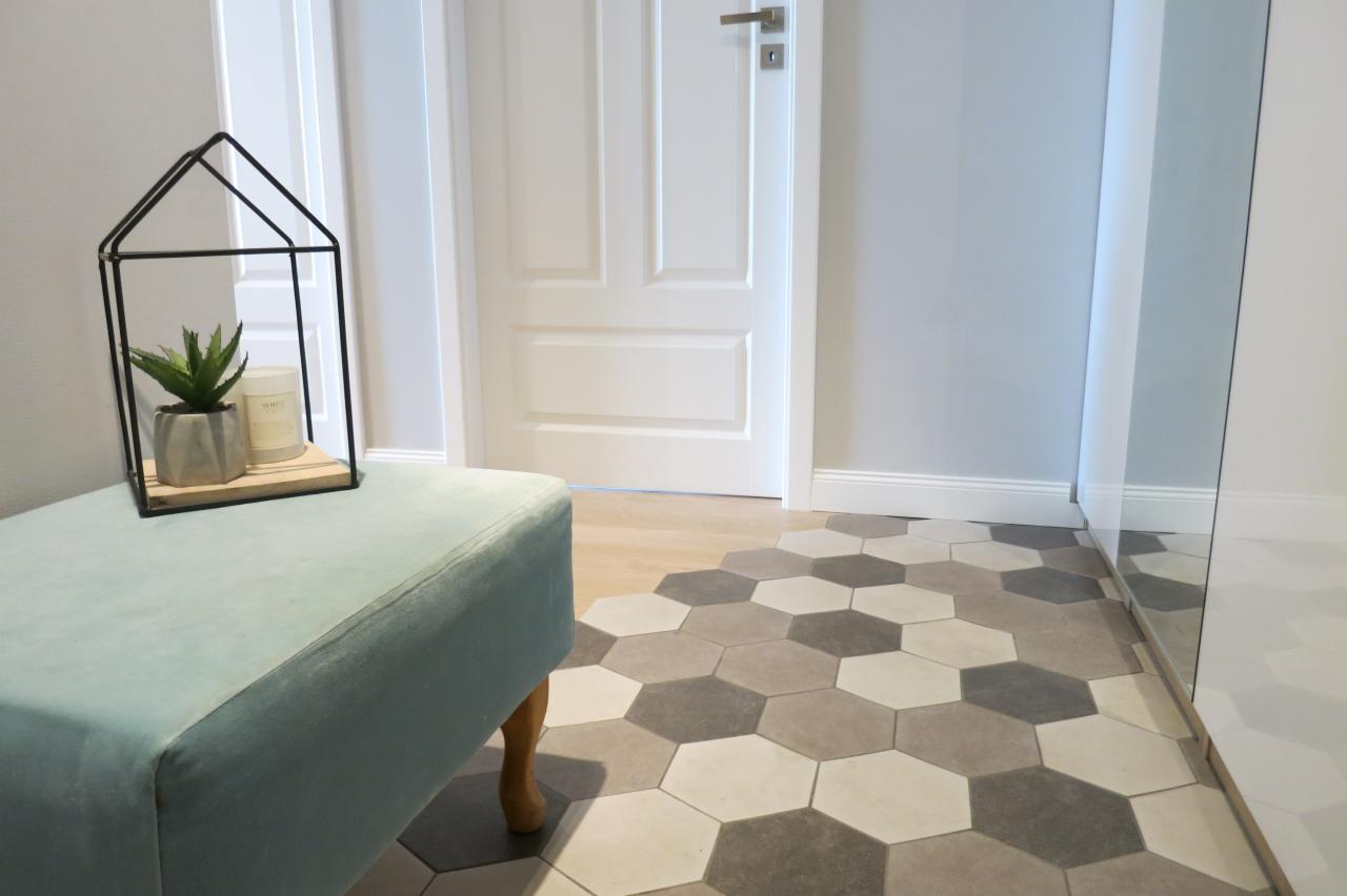 Nowoczesne wnętrze przedpokoju z sześciokątną mozaiką podłogową w odcieniach szarości i beżu, turkusową pufą z drewnianymi nogami oraz dekoracyjną klatką z sukulentem i świecą.