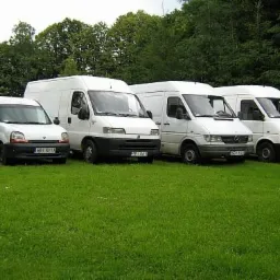 Cztery białe busy różnych marek zaparkowane rzędem na trawniku przed lasem, z widoczną tablicą rejestracyjną pierwszego pojazdu.