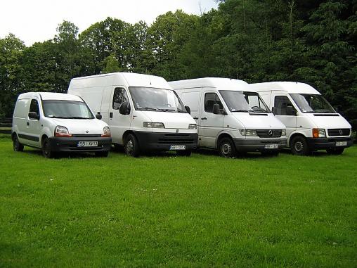 Cztery białe busy różnych marek zaparkowane rzędem na trawniku przed lasem, z widoczną tablicą rejestracyjną pierwszego pojazdu.