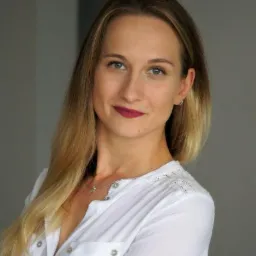 Portret kobiety w białej koszuli z delikatną koronką na ramionach i srebrnym naszyjnikiem, uśmiechającej się lekko do kamery na neutralnym szarym tle.