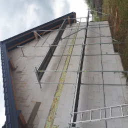 Ściana domu w trakcie murowania z widocznymi pustakami, szalunkami i rusztowaniem, z fragmentem dachu pokrytego czarną dachówką. Widoczne ślady zaprawy i przygotowane miejsca pod ocieplenie.