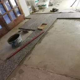 Układanie mozaiki kamiennej na podłodze, widoczny kubeł z klejem, poziomica i narzędzia, w tle puste wiadra i agregat malarski.