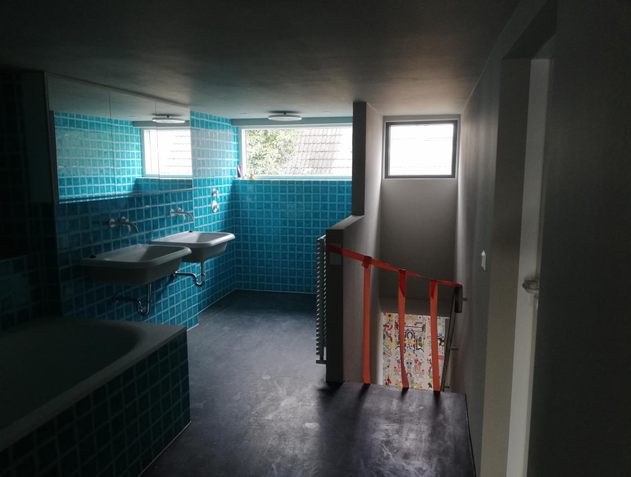 Łazienka z niebieskimi płytkami, dwiema umywalkami na ścianie i wanną, widok na schody zabezpieczone pomarańczową taśmą.