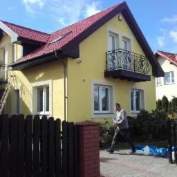 Żółty dom jednorodzinny z czerwonym dachem w trakcie malowania elewacji, pracownik z puszką farby na chodniku, drabina oparta o ścianę, widoczny balkon z ozdobną balustradą.