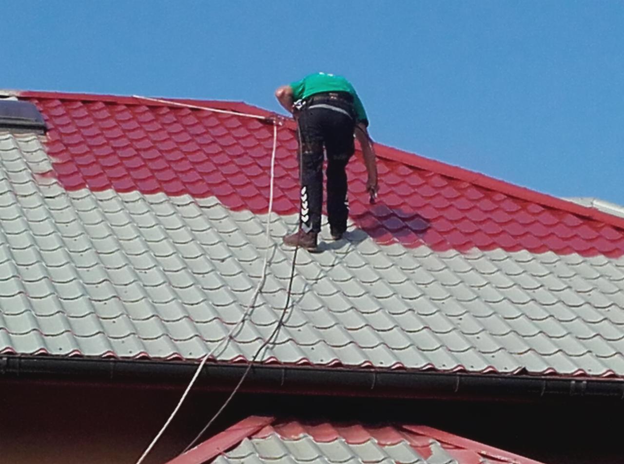 Mężczyzna w uprzęży maluje dachówkę na czerwono, widoczny fragment dachu pomalowany i niepomalowany, biała linka zabezpieczająca.