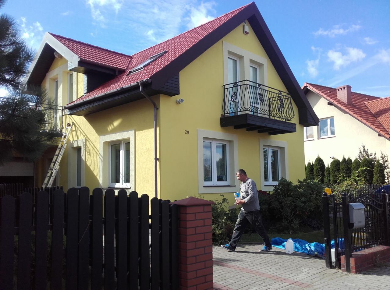 Żółty dom jednorodzinny z czerwonym dachem w trakcie malowania elewacji, pracownik z puszką farby na chodniku, drabina oparta o ścianę, widoczny balkon z ozdobną balustradą.