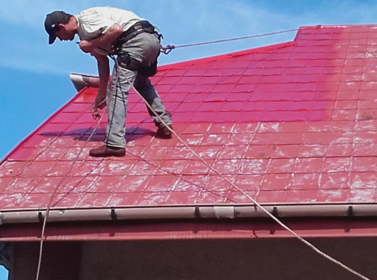 Pracownik zabezpieczony linami maluje czerwony dach z dachówki ceramicznej, widoczne orynnowanie i fragment elewacji budynku.