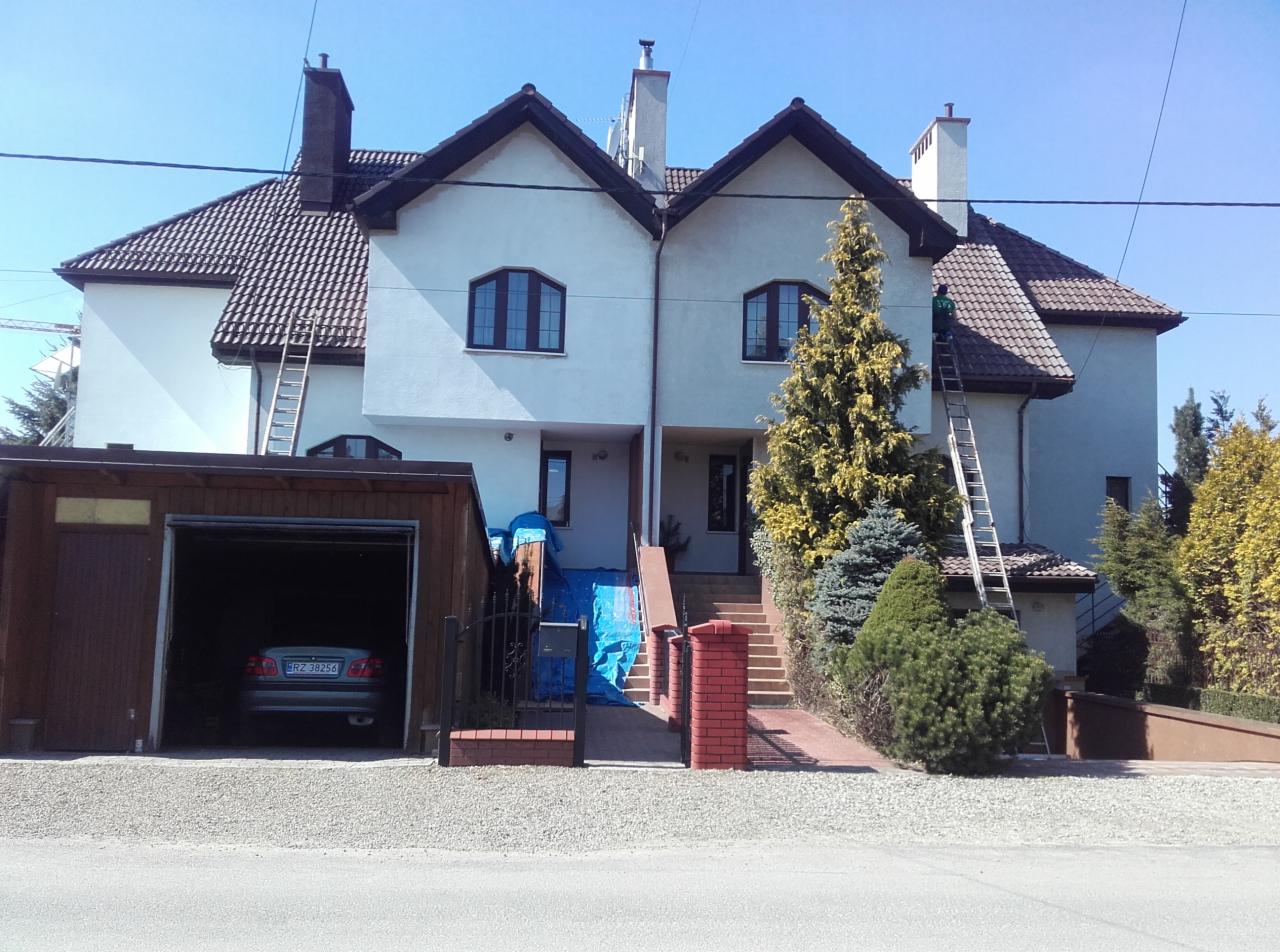 Dwukondygnacyjny dom bliźniak z jasną elewacją w trakcie prac, z rozstawioną drabiną i pracownikiem na wysokości, widoczne zabezpieczenia z folii przy wejściu, garaż z samochodem w tle.