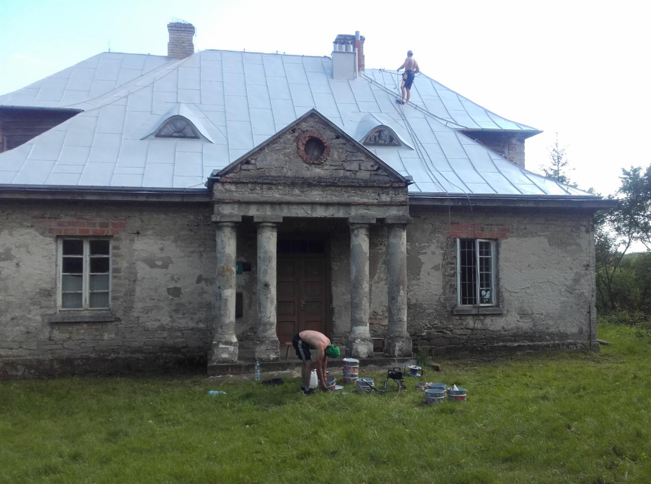 Malowanie dachu zabytkowego dworku z kolumnami; jeden pracownik na dachu, drugi miesza farbę przy puszkach na trawie.