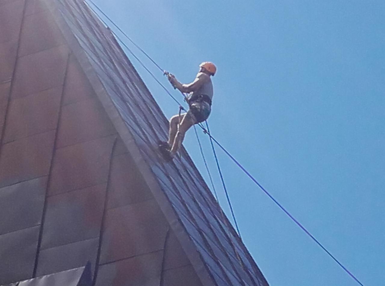 Pracownik na linach w kasku, wykonujący prace na elewacji budynku pokrytej panelami, widok z dołu na tle błękitnego nieba.