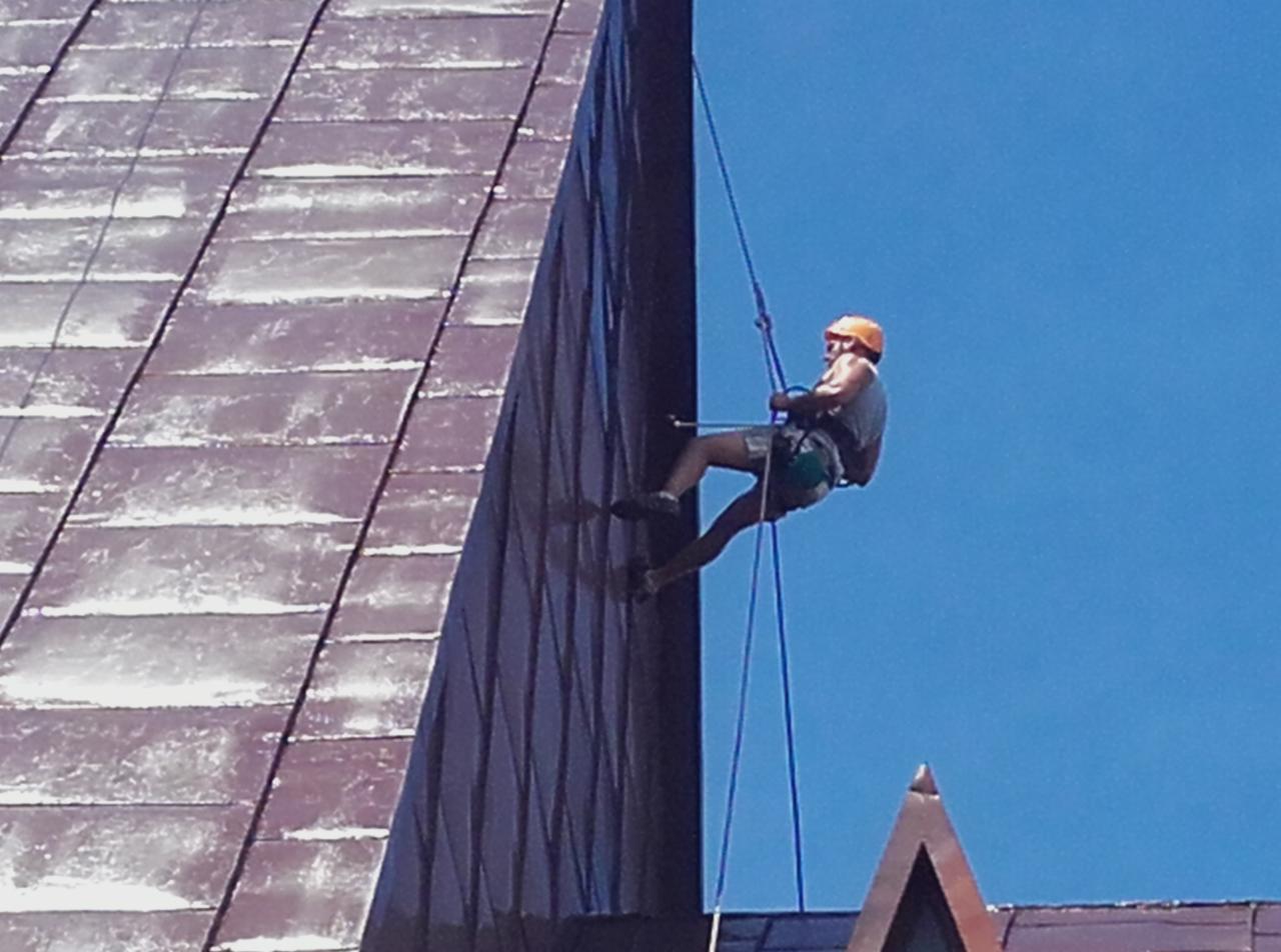Pracownik w kasku, zabezpieczony linami, podczas pracy na wysokości na ciemnej, pionowej ścianie budynku z elementami dachu w tle.