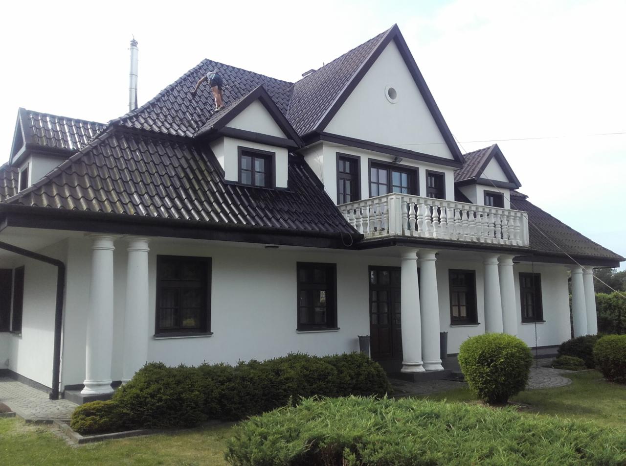 Duży, biały dom z ciemnobrązowym dachem z dachówki ceramicznej, na którym stoi osoba podczas prac konserwacyjnych.