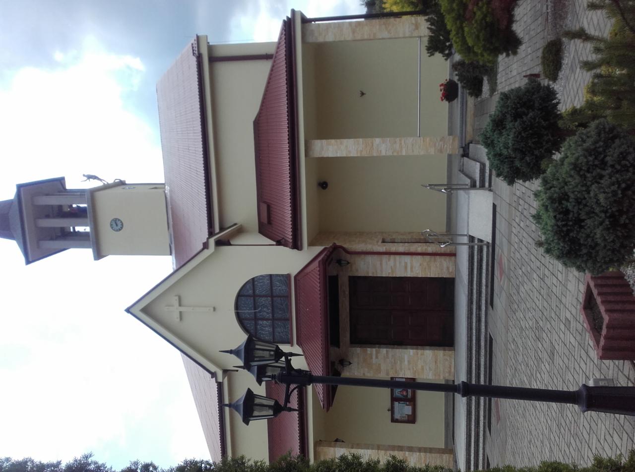 Kościół z beżową elewacją, czerwonym dachem z blachy i wieżą zegarową, widoczny krzyż na fasadzie, oświetlony przez latarnie uliczne.