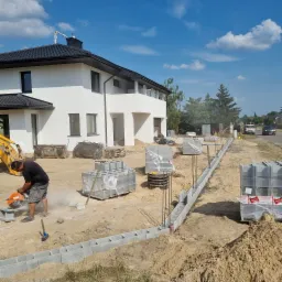 Pracownik przecina szlifierką bloczek betonowy na placu budowy ogrodzenia zbrojonego, w tle nowy dom z białymi ścianami i czarnym dachem.