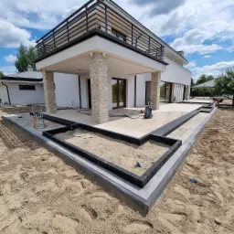 Nowoczesny taras z jasnych płyt i obrzeży z ciemnej kostki, przygotowany pod nasadzenia roślin, z widocznym domem w tle i myjką ciśnieniową.