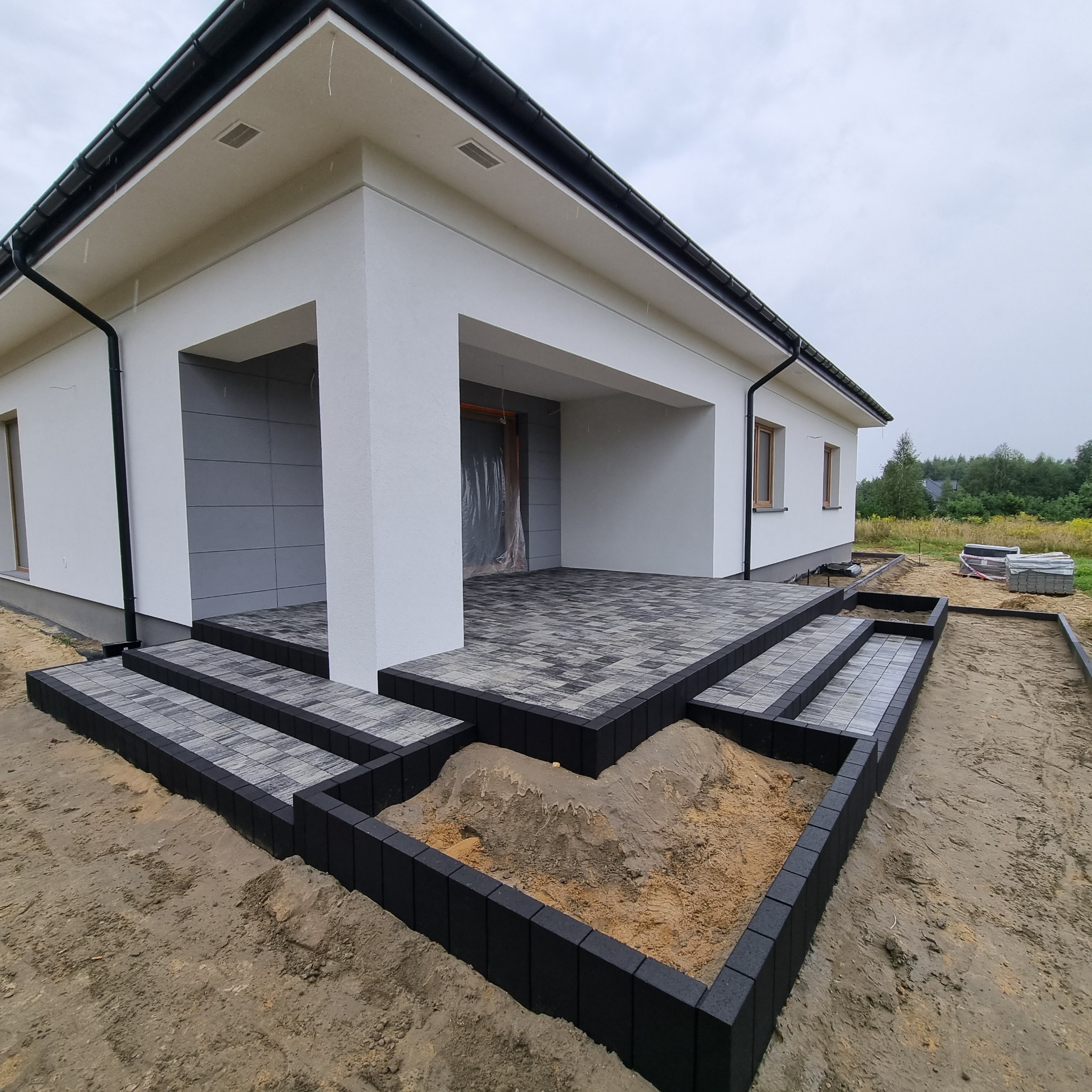 Nowoczesny taras z kostki brukowej w odcieniach szarości, prowadzący do wejścia domu z białymi ścianami i szarą elewacją. Widoczne przygotowane podłoże z obrzeżami, pozostałości piasku i palety...