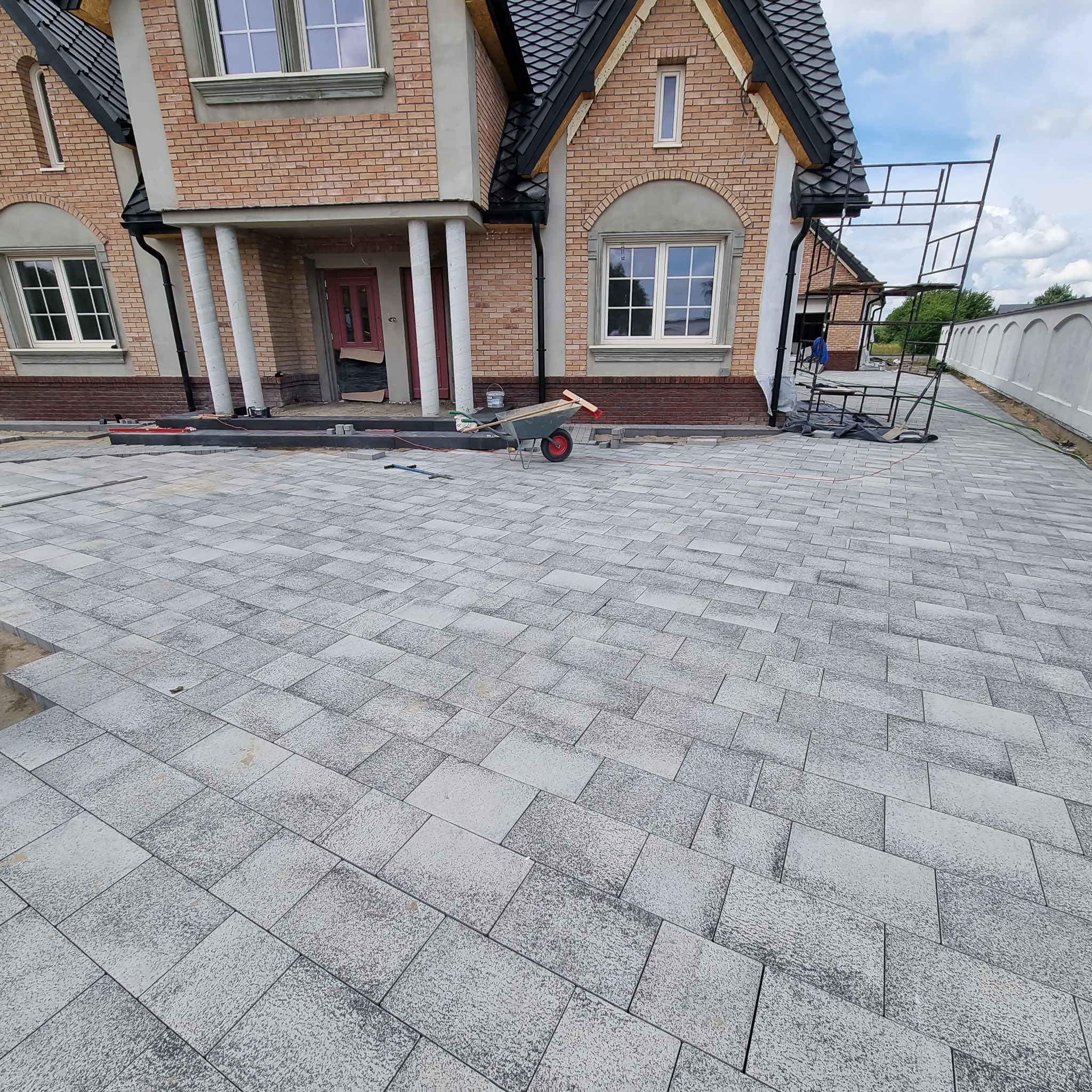 Ułożona szara kostka brukowa przed nowym, dwupiętrowym domem z cegły; widoczny wózek budowlany z narzędziami i rusztowanie przy elewacji.