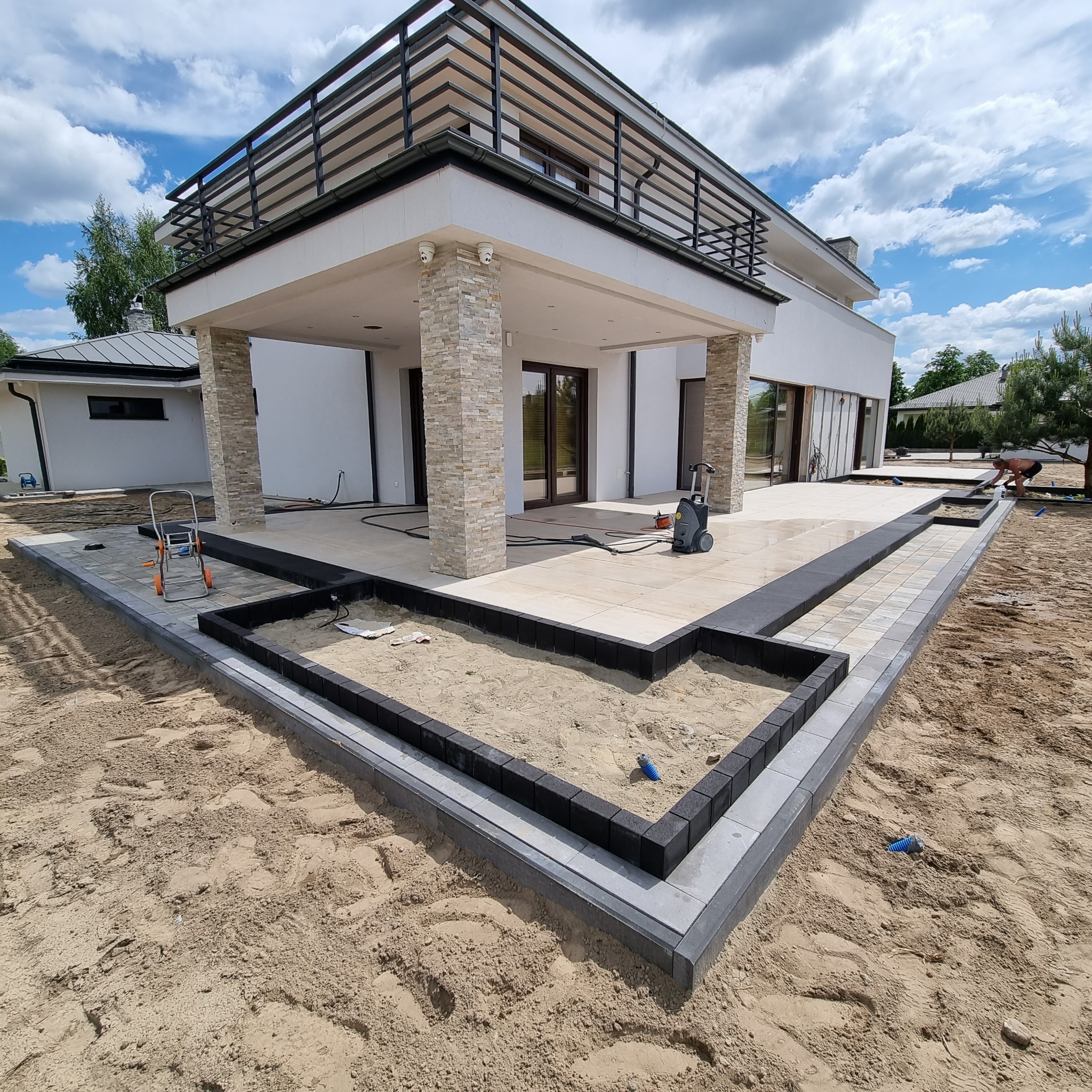 Nowoczesny taras z jasnych płyt i obrzeży z ciemnej kostki, przygotowany pod nasadzenia roślin, z widocznym domem w tle i myjką ciśnieniową.