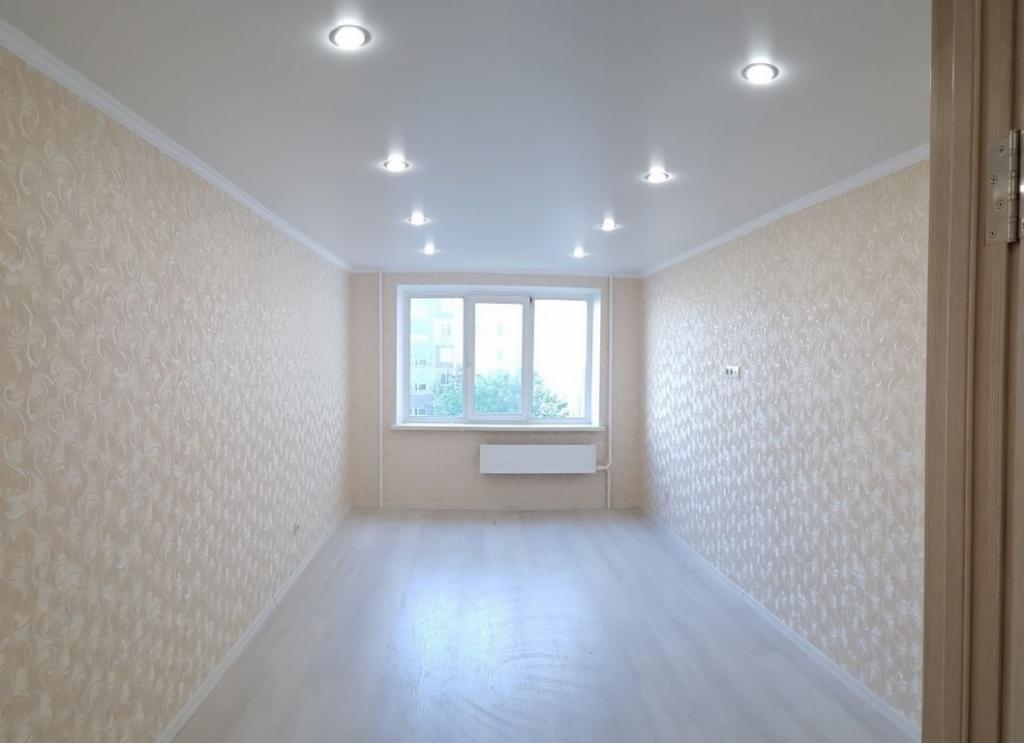 Pusty pokój po remoncie z kremowymi tapetami w ornamenty, jasną podłogą i oświetleniem sufitowym typu downlight, widok na okno z grzejnikiem pod nim. Drzwi po prawej stronie z widocznymi zawiasami.