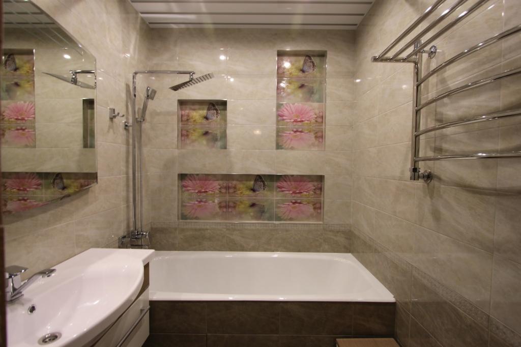 Wykończona łazienka z beżowymi płytkami imitującymi marmur, dekoracyjnymi wnękami z motywem kwiatów i motyli, białą wanną, umywalką z szafką i grzejnikiem drabinkowym.