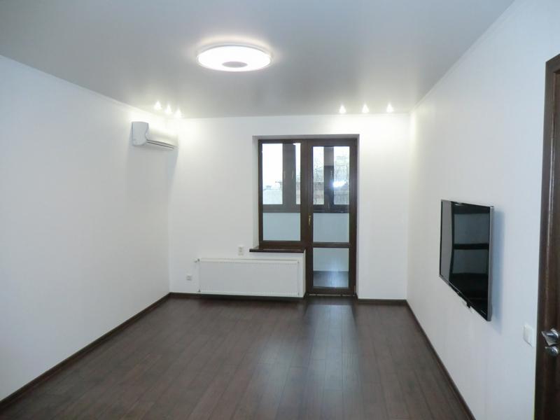 Pusty pokój po remoncie z ciemną podłogą, białymi ścianami, grzejnikiem, klimatyzatorem i telewizorem na ścianie, oświetlony punktowymi lampami i centralnym plafonem.