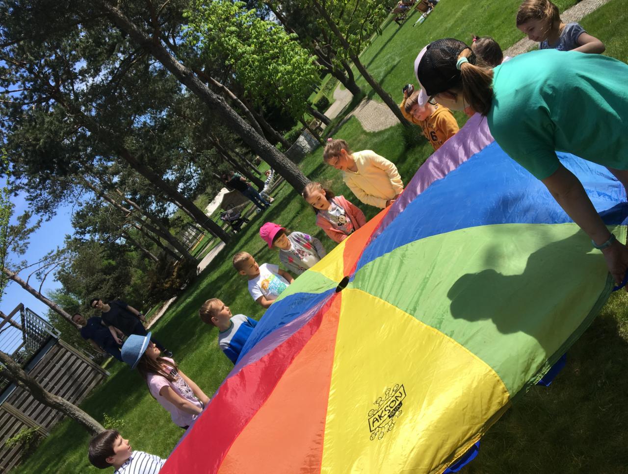Grupa dzieci trzymająca kolorową chustę animacyjną na trawie w parku, widoczne drzewa w tle.