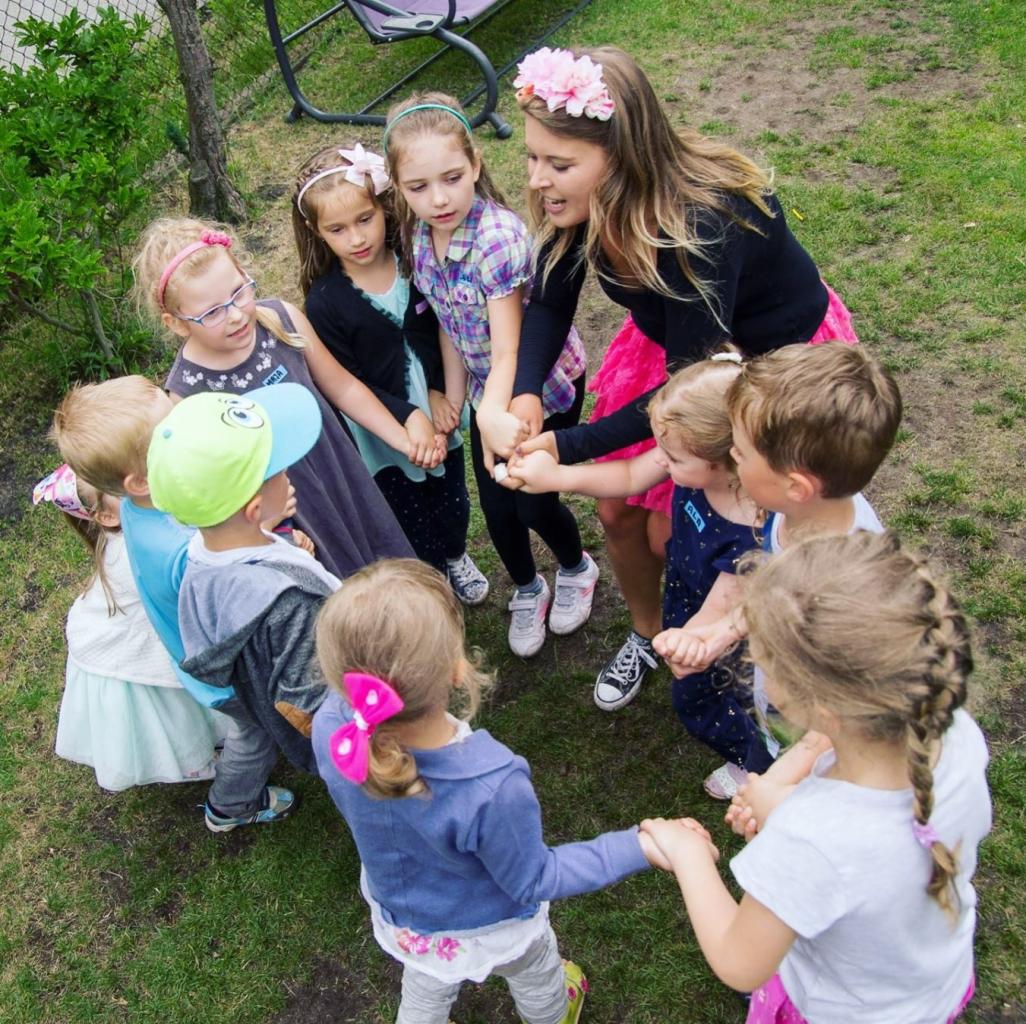 Grupa dzieci trzymających się za ręce na trawie, otoczona przez młodą kobietę z kwiatami we włosach, prawdopodobnie podczas zabawy na świeżym powietrzu.