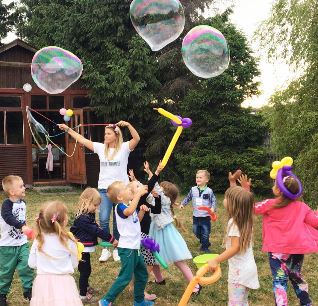 Grupa dzieci bawi się na trawie, próbując złapać duże bańki mydlane puszczane przez kobietę w białej koszulce, w tle drewniany domek i drzewa.