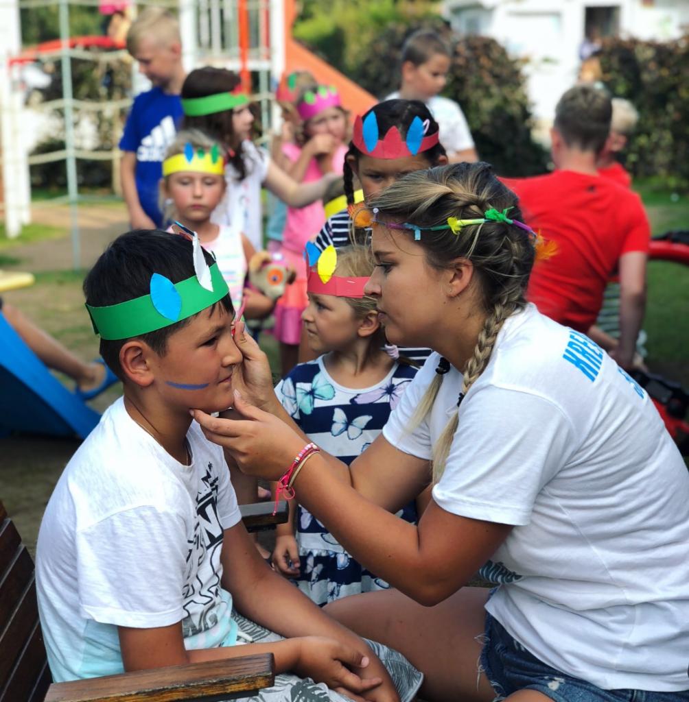 Animator maluje twarz chłopca w indiańskim stroju, w tle grupa dzieci z papierowymi pióropuszami na głowach.