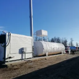 Nowoczesna, biała kotłownia kontenerowa z dużym zbiornikiem na gaz płynny, widoczna na tle błękitnego nieba i zaoranej ziemi, z elementami instalacji zewnętrznej.