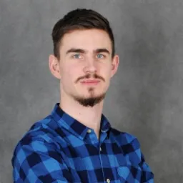 Portret młodego mężczyzny z niebieskimi oczami i zarostem, ubranego w niebieską koszulę w kratę na szarym tle.