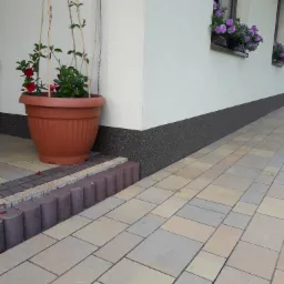 Ułożona kostka brukowa w odcieniach beżu i szarości z dekoracyjnym obrzeżem z brązowych, okrągłych elementów, duża donica z czerwonymi kwiatami przy ścianie budynku, okna z fioletowymi kwiatami w tle.