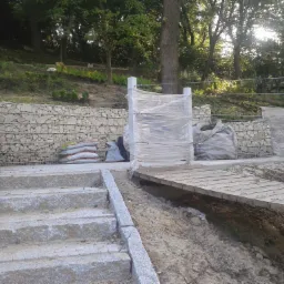 Gabionowe ściany oporowe z kamienia, betonowe schody i drewniany pomost w trakcie budowy na zboczu, zabezpieczone folią i workami z materiałem budowlanym, z widokiem na zalesiony teren.