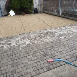 Fragment podjazdu z częściowo ułożoną szarą kostką brukową i wysypanym żwirem, widoczna taczka z żwirem i zielony krzew w tle.