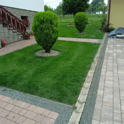 Utrzymany trawnik z dwoma formowanymi krzewami, obrzeża z kostki brukowej i kamienia, dom z kamienną elewacją i drewnianymi schodami w tle.