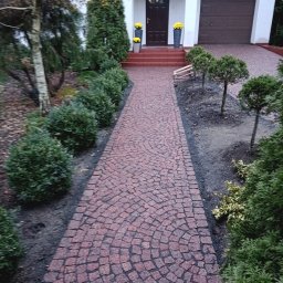 MONUMENTICA Andrzej Lenski - Czerwona kostka granitowa ułożona w łukowaty wzór tworzy ścieżkę do domu z numerem 58. Po bokach ścieżki rosną krzewy i drzewka, a na schodach stoją donice z żółtymi kwiatami.