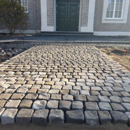 MONUMENTICA Andrzej Lenski - Ścieżka z kostki granitowej prowadząca do wejścia domu z szarej cegły. Częściowo ułożona kostka, widoczne przygotowania do dalszych prac. Jasne, naturalne światło.