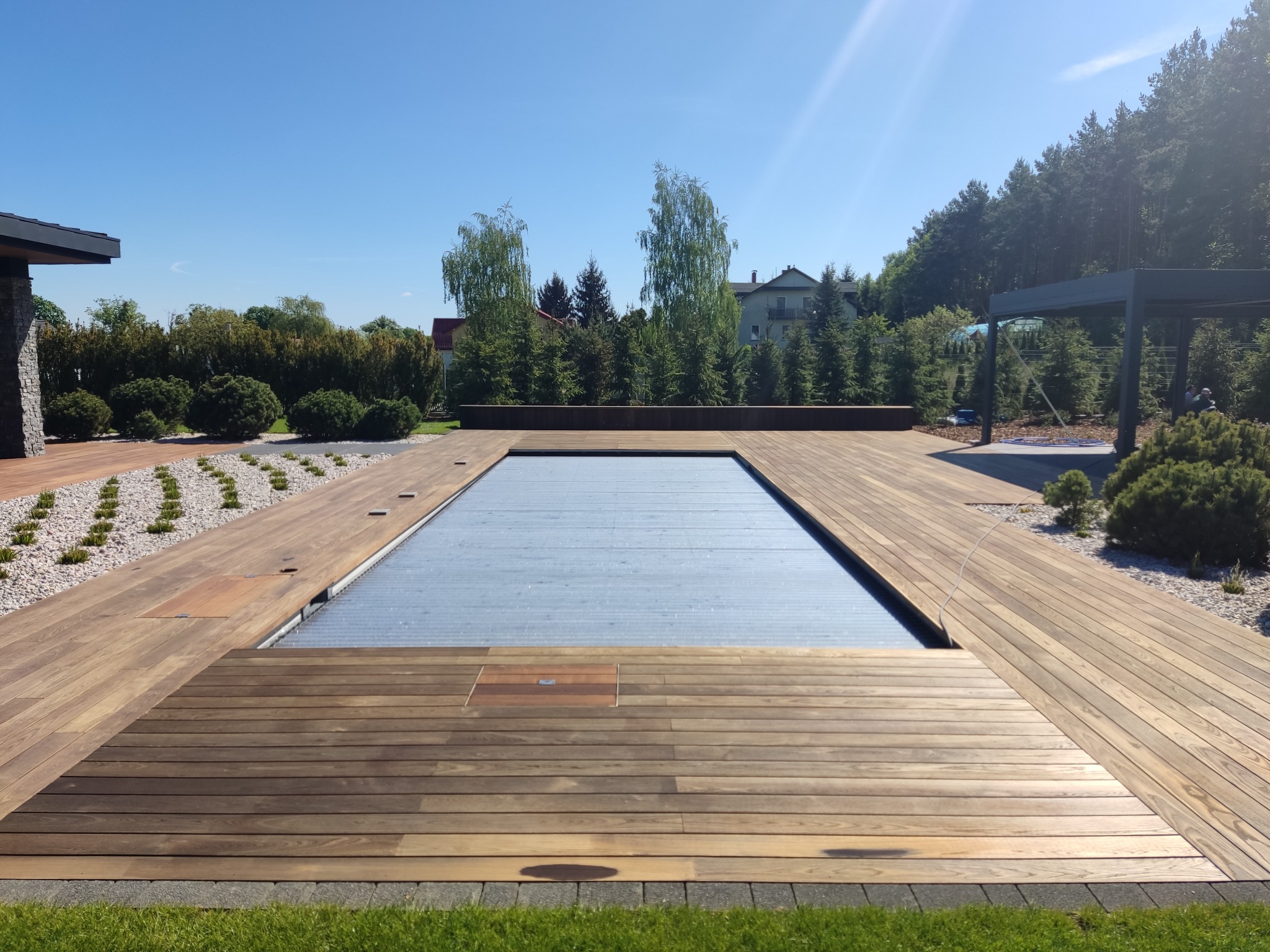 Nowoczesny taras z drewna otaczający basen z automatyczną roletą. Widok na ogród z drzewami i błękitne niebo w słoneczny dzień. Estetyczne wykończenie.