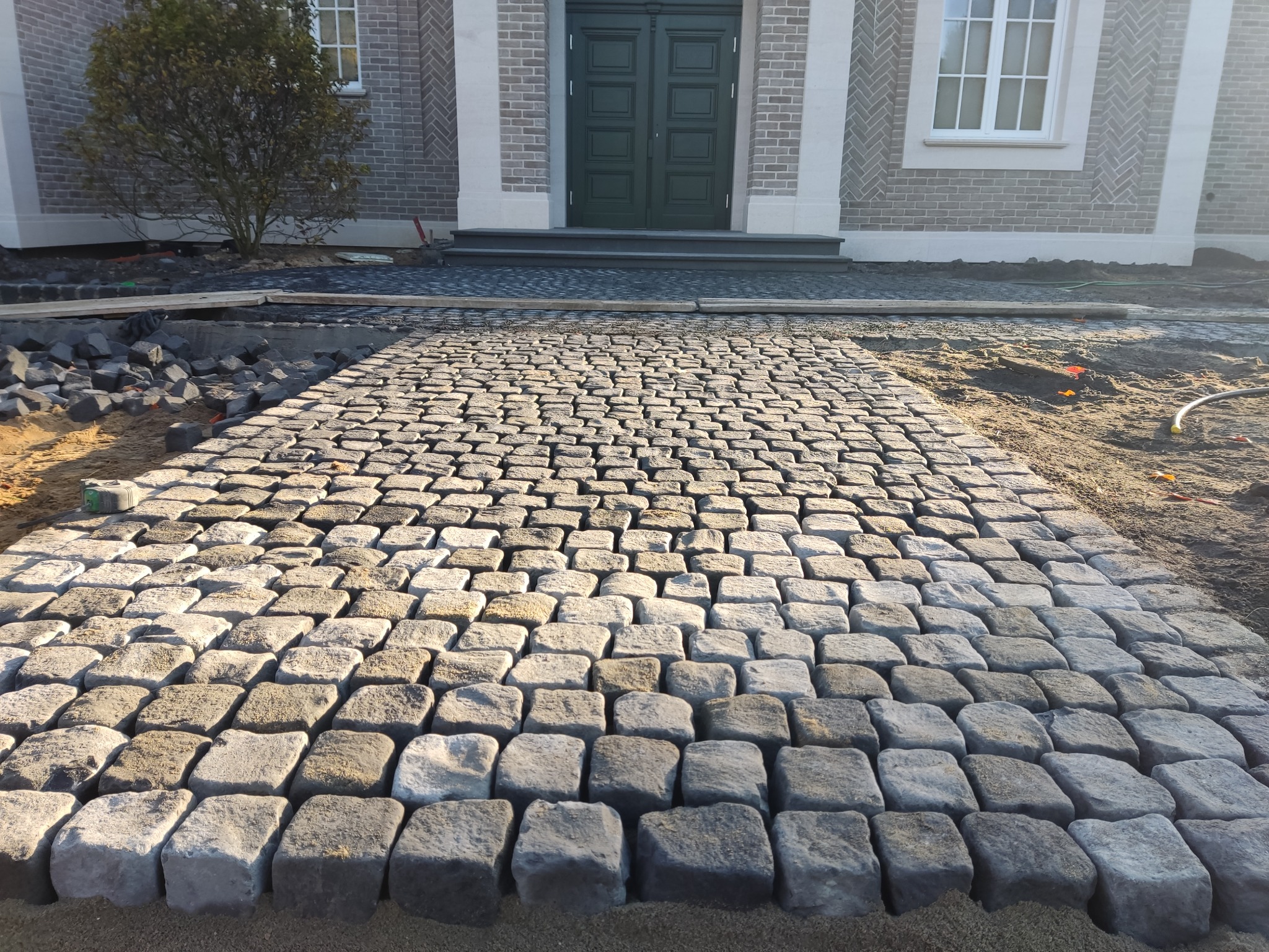 Ścieżka z kostki granitowej prowadząca do wejścia domu z szarej cegły. Częściowo ułożona kostka, widoczne przygotowania do dalszych prac. Jasne, naturalne światło.