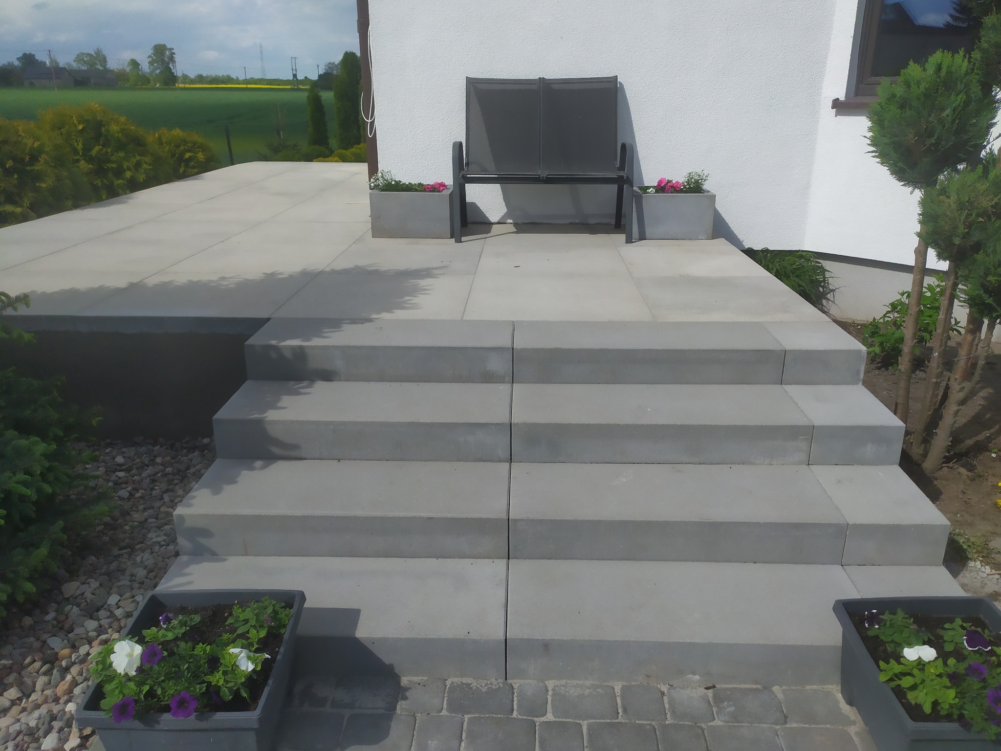 Nowoczesne schody z szarej kostki brukowej prowadzące do tarasu z ławką i kwiatami w donicach. Widok na zielone pola w tle.