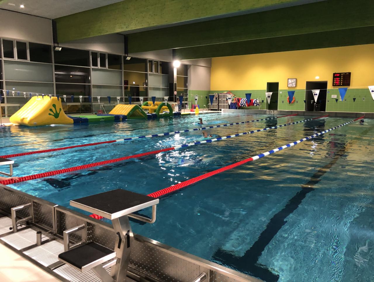 Kryty basen z torami pływackimi, dmuchanym torem przeszkód dla dzieci i drabinkami startowymi. W tle widoczne okna i ściana z zegarem.