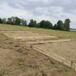 Przygotowanie terenu pod fundamenty: drewniane szalunki ułożone na trawiastym podłożu, widoczne wiązania i poziomice, w tle drzewa i pochmurne niebo.