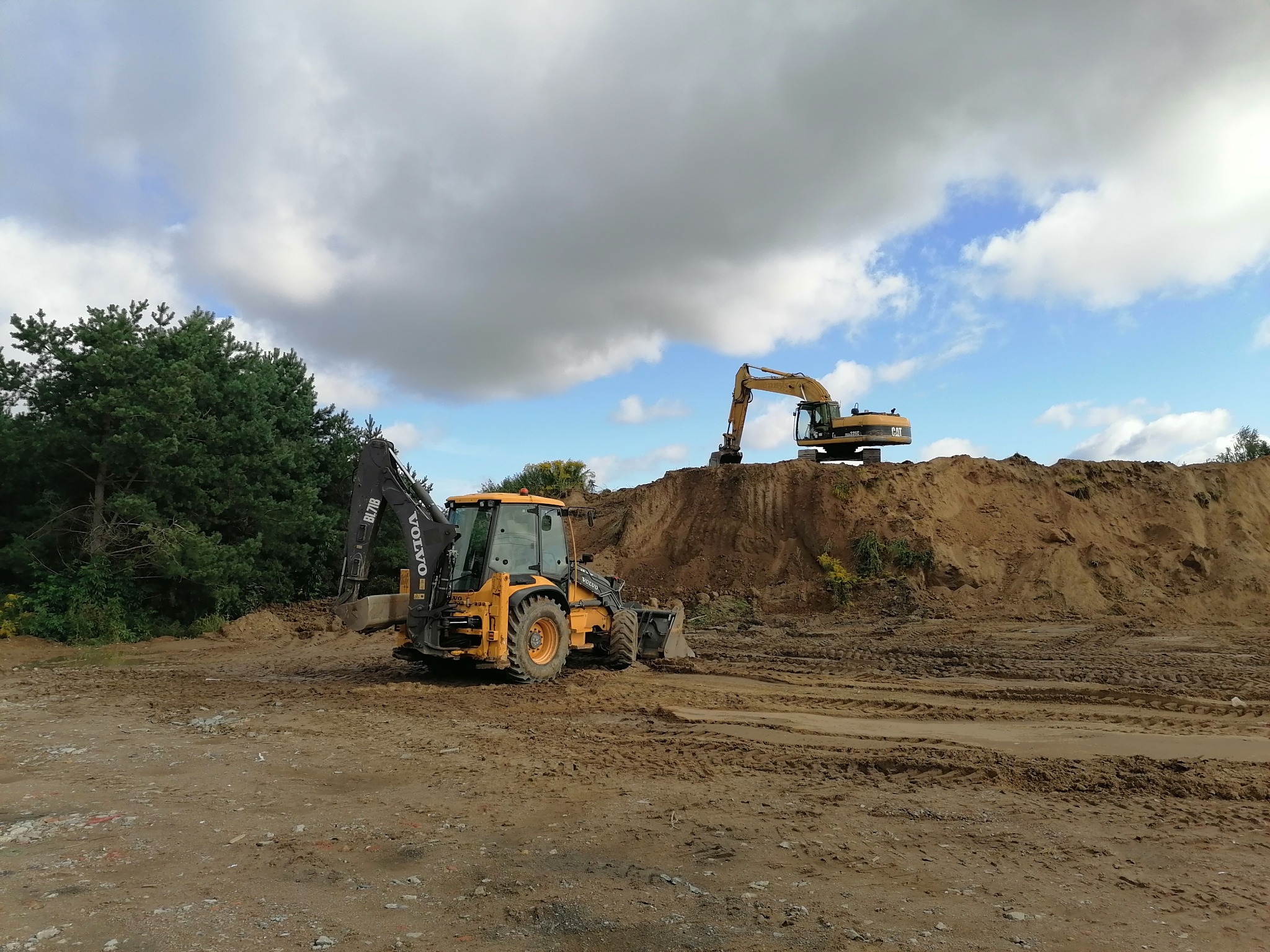 Żółta koparko-ładowarka Volvo i koparka Caterpillar na placu budowy z dużą pryzmą ziemi, widoczne ślady opon, zieleń drzew w tle i pochmurne niebo.