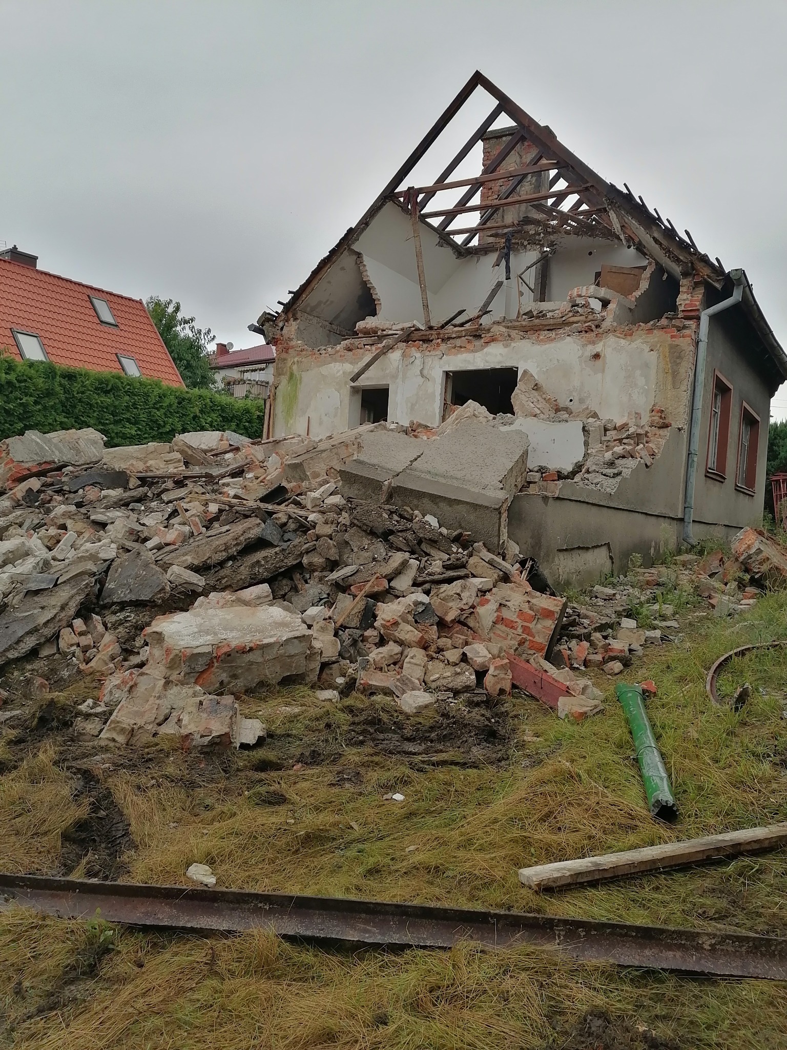 Zdewastowany budynek z częściowo zawalonym dachem, gruzowisko z cegieł i betonu na trawiastym terenie, widoczne elementy konstrukcji dachu i fragmenty ścian.