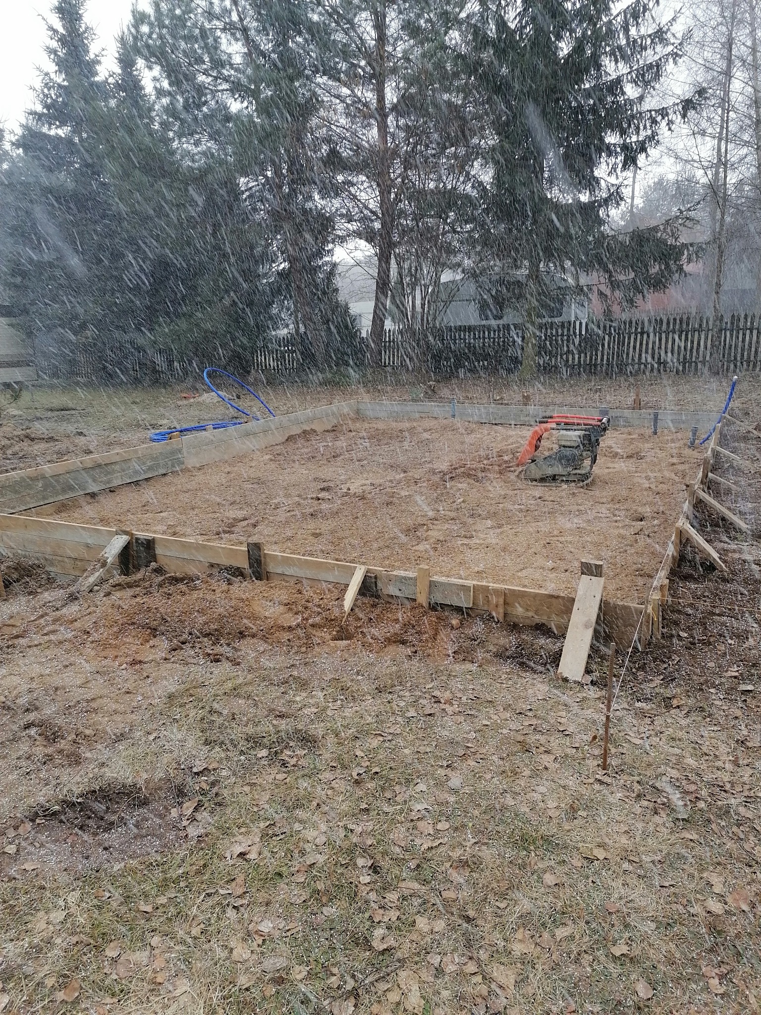 Przygotowanie terenu pod fundamenty w trakcie opadów śniegu. Widoczny szalunek z desek, ubijarka mechaniczna, rury doprowadzające wodę i zasypka piaskowa.