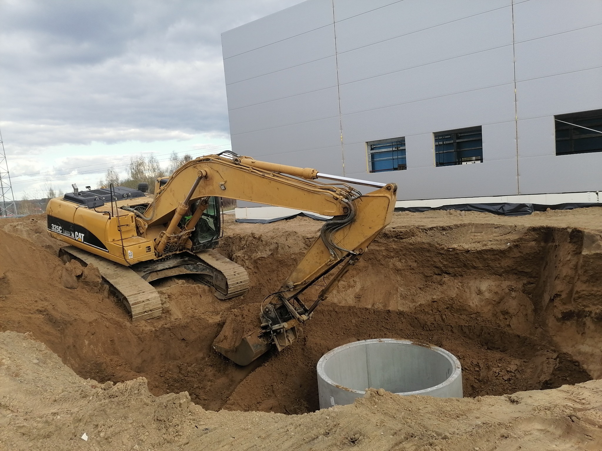 Żółta koparka CAT 325C w trakcie wykopu ziemi obok betonowego kręgu na tle szarej hali i pochmurnego nieba.