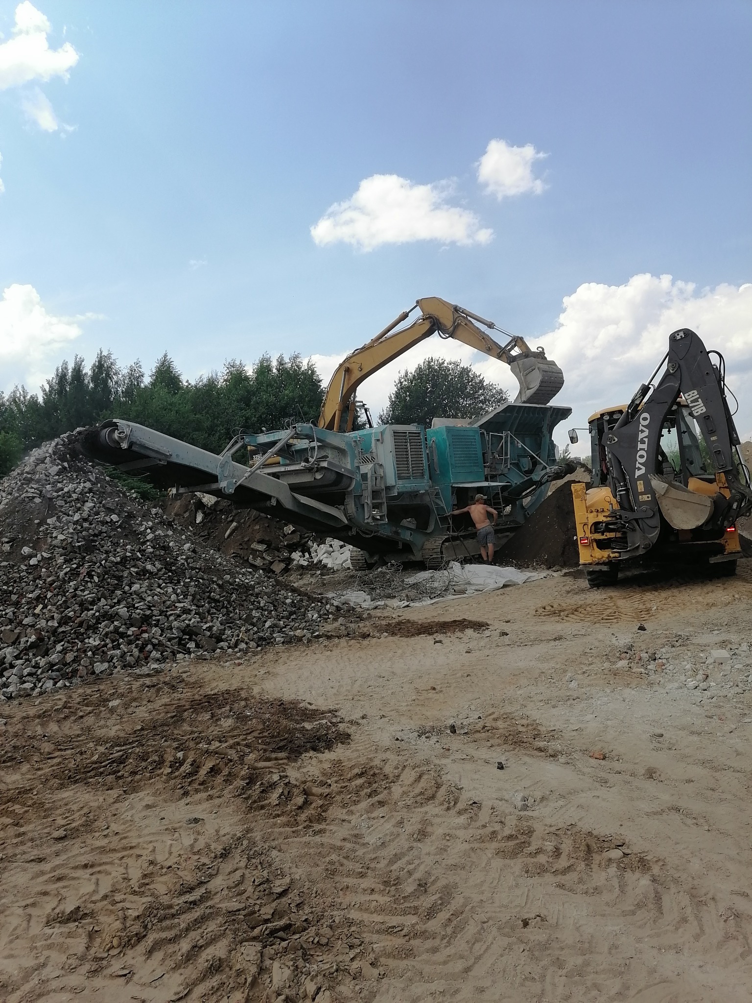 Praca kruszarki do gruzu na placu budowy z operatorem i koparką Volvo, widoczne ślady opon na piasku i duża góra pokruszonego materiału.