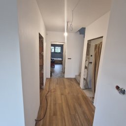 Tom-Fliz - Korytarz w trakcie prac wykończeniowych: świeżo pomalowane ściany na biało, drewnopodobna podłoga, widoczne instalacje elektryczne i otwory drzwiowe bez ościeżnic.