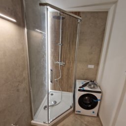 Tom-Fliz - Nowoczesna kabina prysznicowa z deszczownicą w wykończonej łazience, obok pralka. Ściany wyłożone płytkami imitującymi drewno. Minimalistyczny design.